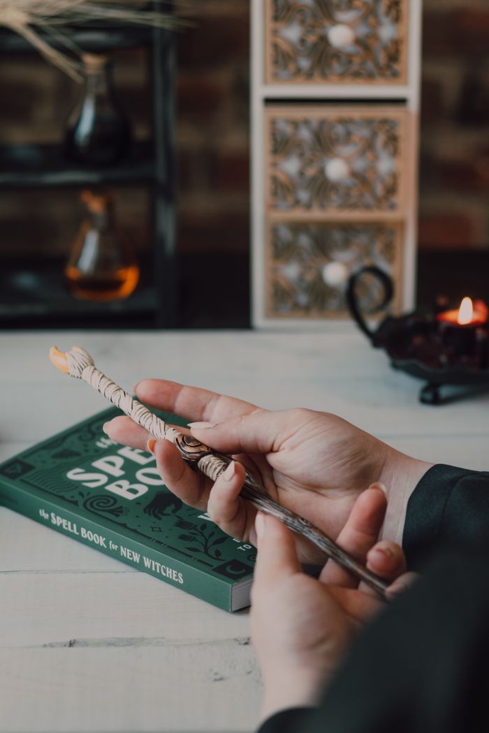 Close-up of hands holding a wand with a spell book on a wooden table indoors.